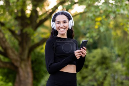 Photo for Active brunette woman wearing wireless headset and black sportswear while looking at camera with smartphone in hand. Smiling female doing daily exercises to music on background of public park. - Royalty Free Image