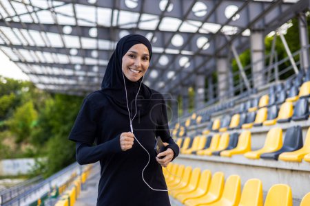 Mujer islámica positiva en hijab y ropa deportiva haciendo ejercicios con auriculares con cable conectados al teléfono móvil. Sonriente dama disfrutando de pasar tiempo en el estadio y practicar correr a la música.