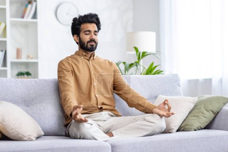 Tranquilo hombre de Oriente Medio sentado en un sofá almohadillado en posición siddhasana y usando las manos mudra con los ojos cerrados. Tranquilo chico respirando profundamente y practicando yoga en casa para reducir los niveles de estrés.