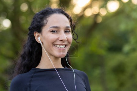 Optimistic diverse sportswoman in black top smiling at camera while listening to motivating playlist in wired earphones. Attractive female in black top finishing training in green public area.