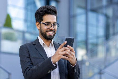 An Indian businessman in formal wear is engaged with his smartphone in front of a contemporary office setting, depicting connectivity and corporate lifestyle.