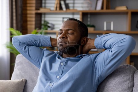 Heiterer afroamerikanischer Mann mit geschlossenen Augen, der die Ruhe genießt, während er auf einem Sofa in einer gemütlichen Wohnzimmeratmosphäre sitzt.