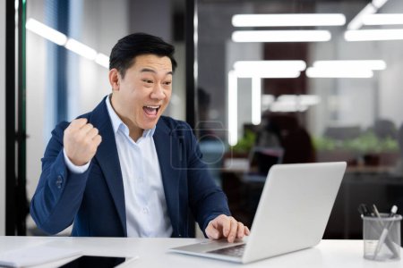 Téléchargez les photos : Entrepreneur masculin excité exprimant sa joie pour son succès dans un cadre d'entreprise, avec un ordinateur portable dans un bureau lumineux. - en image libre de droit