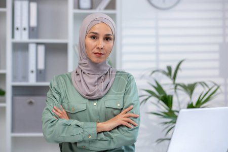 Professional young Muslim woman wearing a hijab stands confidently with arms crossed in a bright office environment.