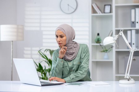 A dedicated professional wearing a hijab is deeply focused as she works on her laptop at a bright office desk.