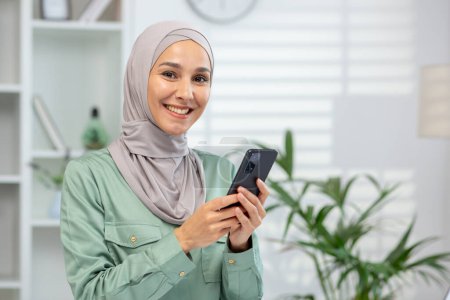 Portrait of a cheerful Muslim woman wearing a hijab, holding a phone, standing in a cozy home setting with plants.