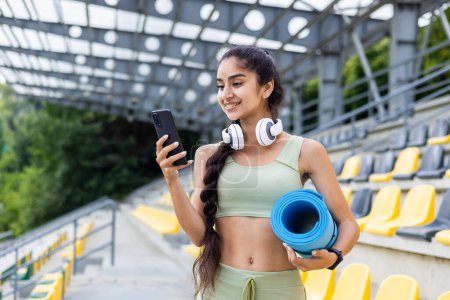 Active young Indian woman with headphones and yoga mat at stadium using smartphone, preparing for her daily fitness routine.