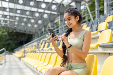 Inderin im Trainingsanzug überprüft Fitnessarmband und Smartphone während Trainingspause im Stadion und zeigt aktiven Lebensstil.