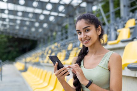 Téléchargez les photos : Joyeux jeune femme indienne synchronisation smartwatch fitness avec téléphone au stade pendant le jogging de jour et séance d'entraînement. - en image libre de droit