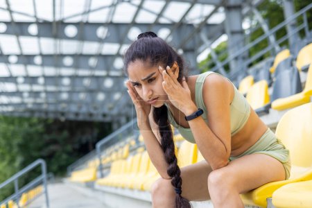 Foto de Mujer india agotada en un chándal tomando un descanso durante una rutina de fitness en un estadio, revisando su reloj inteligente. - Imagen libre de derechos