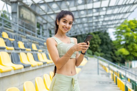 Téléchargez les photos : Femme indienne souriante en vêtements de sport avec smartwatch, en utilisant le téléphone au stade. Concept de mode de vie actif et de technologie dans le sport. - en image libre de droit