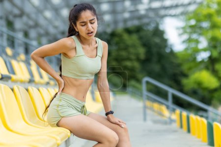 Foto de La joven india enfocada en chándal detiene su rutina de jogging en un estadio, mostrando su pulsera de fitness. - Imagen libre de derechos