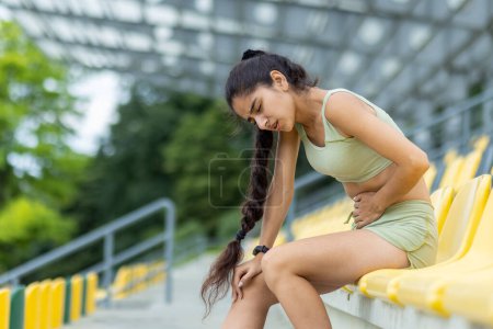 Foto de Mujer india joven en chándal con reloj inteligente descansando durante la sesión de jogging al aire libre en un estadio, encarnando un estilo de vida saludable. - Imagen libre de derechos