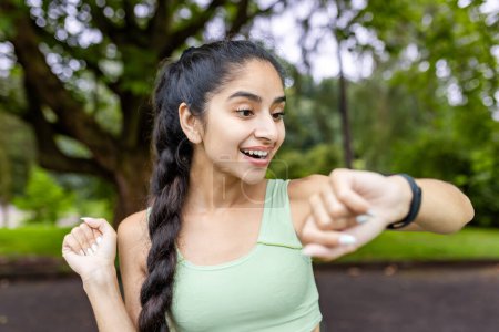 Motivated Indian woman in a tracksuit uses a fitness tracker while running in a lush green park, embodying health and vitality.
