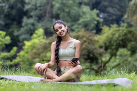 A young Indian woman in a tracksuit sits on a yoga mat, smiling serenely amidst green trees in a tranquil park.