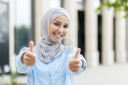 Eine fröhliche junge Frau im Hijab streckt mit einem breiten, selbstbewussten Lächeln einen doppelten Daumen in die Höhe und signalisiert Zustimmung und Erfolg.