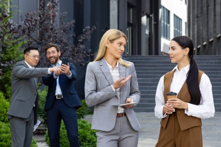Bullying, sexism and ridicule in the team at work. Young women are standing talking to themselves, businessmen are standing behind and laughing, looking at the phone screen.