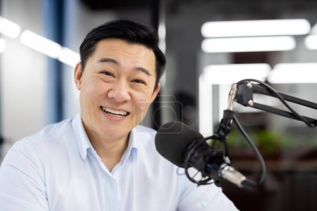 Photo for Close-up portrait of a young Asian man sitting in the office in front of a microphone and smiling at the camera. - Royalty Free Image
