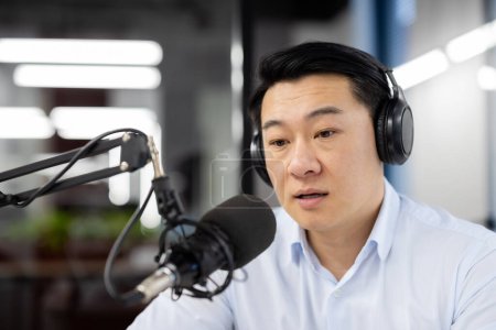 Photo for Asian young man sitting in office wearing headphones and in front of desk with microphone, talking remotely with clients, recording business training. Close-up photo. - Royalty Free Image