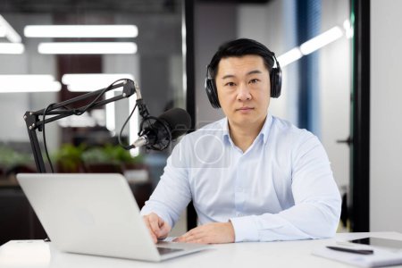 Photo for Portrait of a serious young businessman sitting in the office at the desk wearing headphones and in front of a laptop, confidently looking at the camera. - Royalty Free Image