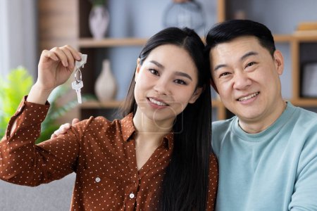 Sonriente pareja asiática mostrando una llave de la casa, que representa la nueva propiedad de la vivienda y la felicidad en su acogedor ambiente hogareño.