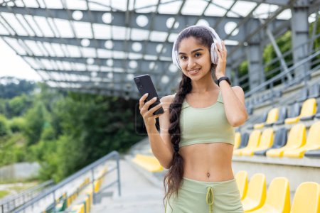 Foto de Retrato de una joven y sonriente chica india deportiva de pie en el estadio tribuna con auriculares, sosteniendo un teléfono y mirando a la cámara. - Imagen libre de derechos