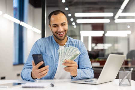 Happy businessman in a casual blue shirt holding cash and a mobile phone, feeling successful at work in a modern office.