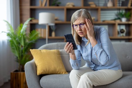 An elderly woman sits on a couch, expressing confusion while looking at her smartphone, possibly dealing with technology issues.