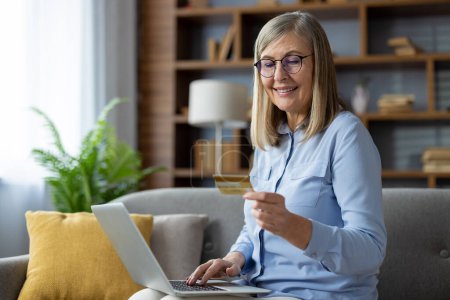 Téléchargez les photos : Femme mûre avec des lunettes sourit comme elle utilise un ordinateur portable pour faire un achat en ligne en utilisant une carte de crédit dans un cadre confortable à la maison. - en image libre de droit