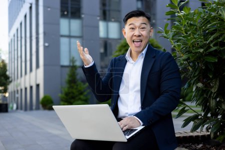 Téléchargez les photos : Homme d'affaires joyeux avec ordinateur portable assis à l'extérieur, geste triomphant dans un cadre urbain moderne. - en image libre de droit