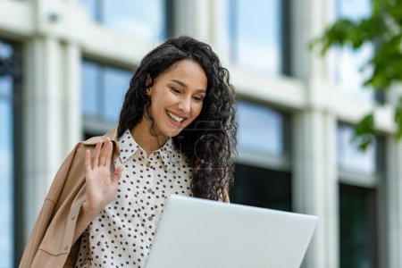 Una mujer de negocios confiada con un ordenador portátil está saludando fuera de un moderno edificio de oficinas, encarnando la elegancia profesional y la amabilidad.