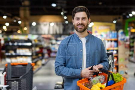 Un macho adulto sostiene una cesta de compras con productos frescos mientras compra en la sección de comestibles de un supermercado..