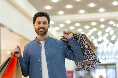 Photo for A cheerful man carrying shopping bags, smiling at the camera in a brightly lit mall setting, giving a sense of retail therapy and consumerism. - Royalty Free Image