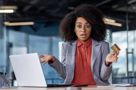Schockierte Afroamerikanerin, die am Schreibtisch mit Laptop die Hände mit Kreditkarte spreizt. Verwirrter Büroangestellter kämpft mit Online-Transaktion wegen schwacher Verbindung im Kabinett.