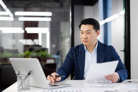 Un hombre de negocios profesional con un traje azul revisa los informes mientras trabaja en su computadora portátil en un entorno de oficina moderno.
