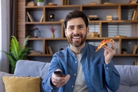 Fröhlicher Mann mittleren Alters mit Pizzascheibe und Fernbedienung, der es sich auf der Couch in seinem gemütlichen Wohnzimmer gemütlich macht, mit glücklichem Gesichtsausdruck.
