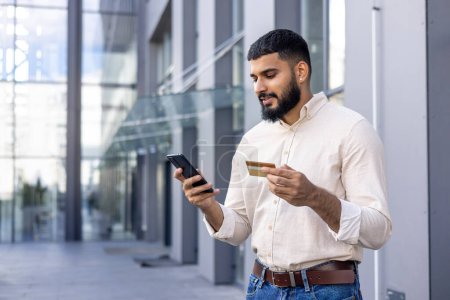Selbstbewusster junger erwachsener Mann mit Smartphone und Kreditkarte an einem sonnigen Tag vor modernen Bürogebäuden. Konzept von Mobile Banking, E-Commerce und modernem Lifestyle.