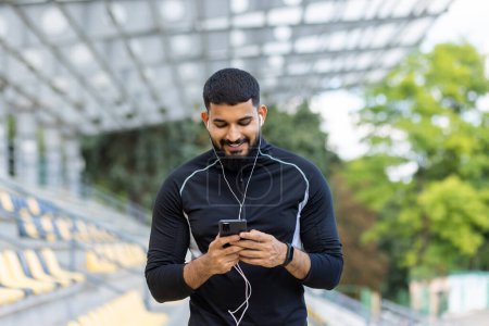 A young, athletic man in sportswear is smiling while using his smartphone and wired earphones at a stadium, conveying a sense of leisure and technology.
