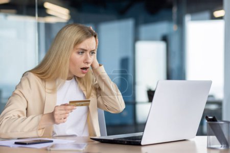 Businesswoman in office experiencing shock while holding a credit card and staring at a laptop screen, likely encountering unexpected charges.