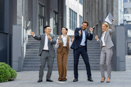 Cuatro colegas profesionales de negocios celebran alegremente una victoria con las manos levantadas fuera de un edificio moderno.