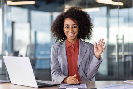 Eine nahbare afroamerikanische Geschäftsfrau winkt fröhlich, während sie an ihrem Büroarbeitsplatz sitzt und Freundlichkeit und Professionalität verkörpert.