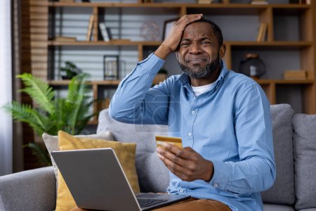 Photo for Adult male at home looking at his laptop with a concerned expression, holding a credit card, potentially a victim of online fraud. - Royalty Free Image