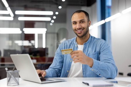 Un homme souriant tenant une carte de crédit tout en utilisant un ordinateur portable, représentant les transactions en ligne sécurisées et la banque numérique.