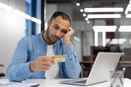 Un hombre parece preocupado mientras sostiene una tarjeta de crédito y mira la pantalla de su computadora portátil, posiblemente lidiando con problemas financieros.