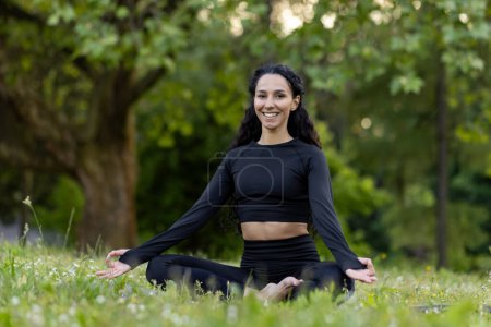 Photo for A serene young woman in black sportswear meditating in a lotus pose amidst vibrant green foliage and a calm, natural setting. - Royalty Free Image