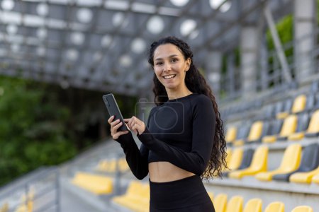 Eine lächelnde junge Frau in Sportkleidung hält ein Smartphone in der Hand, während sie an einem Sportstadion steht, im Hintergrund sitzen Sitze..
