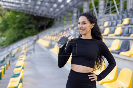 A fitness enthusiast holding a water bottle, taking a break during an outdoor workout session in a stadium.