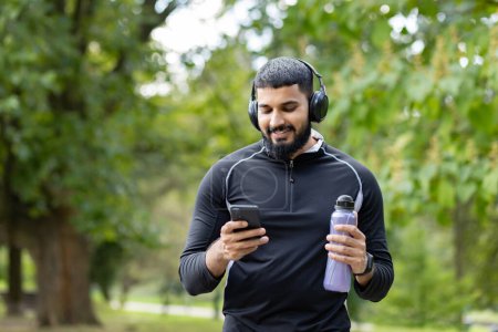 Smiling man in sportswear holding a water bottle while using his smartphone and wearing headphones in a park setting.