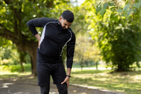 An athletic man in sportswear holds his lower back in pain while standing in a lush green park, showing signs of injury.