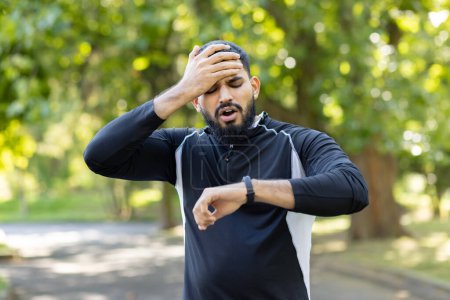 A man in workout clothes appears confused checking his smartwatch in the park, possibly having missed his exercise routine.